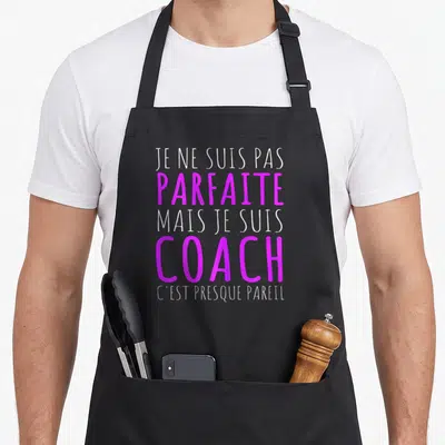 Tablier de cuisine noir avec le texte Je ne suis pas parfaite mais je suis coach en lettres blanches et violettes.