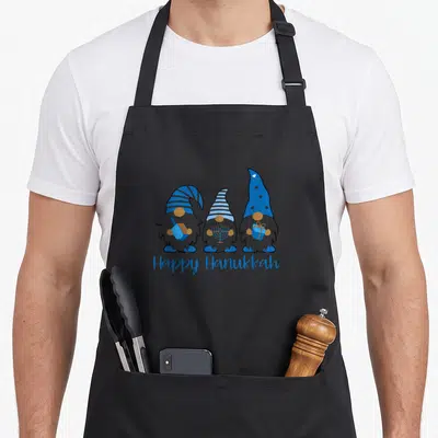Tablier de cuisine noir avec trois nains de Hanoucca bleus et gris tenant un dreidel, une menorah et un cadeau sous linscription Happy Hanukkah.
