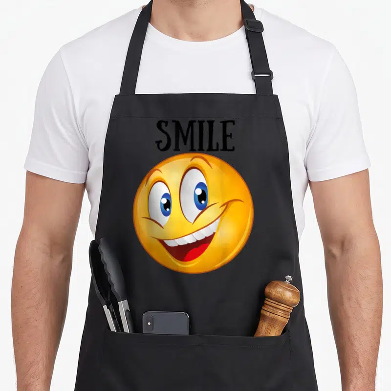 Tablier de cuisine noir avec un grand smiley jaune souriant et le mot SMILE en lettres noires au-dessus.