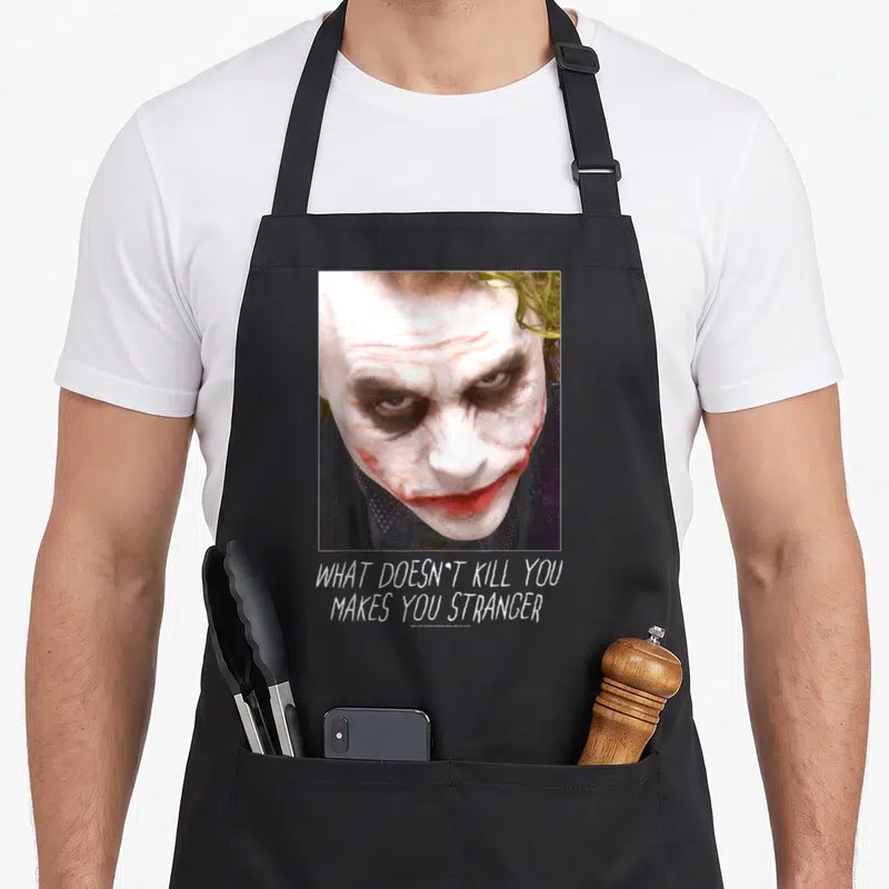 Tablier de cuisine noir avec le visage du Joker et la phrase What Doesn't Kill You Makes You Strangerer imprimée en blanc.