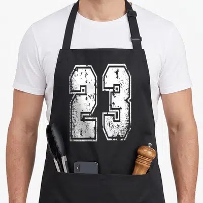 Tablier de cuisine noir avec le numéro 23 imprimé en blanc dans un style maillot de sport délavé et vintage.