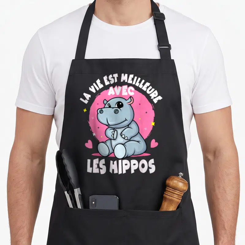 Tablier de cuisine noir avec un hippopotame gris assis devant un cercle rose et des étoiles jaunes.