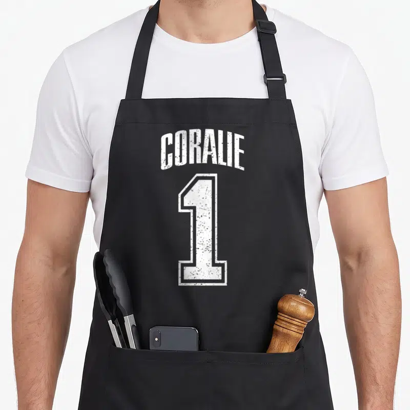 Tablier de cuisine noir avec le nom Coralie et le chiffre 1 blanc en typographie sportive vintage sur le devant.