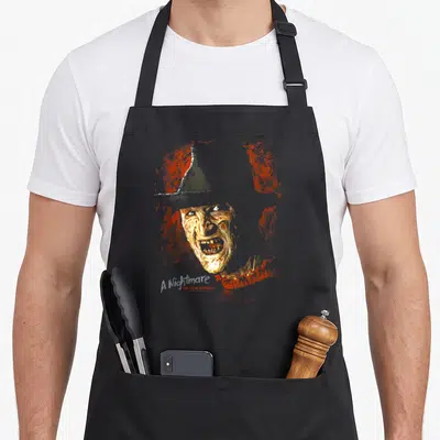 Tablier de cuisine noir avec portrait stylisé de Freddy Krueger et texte A Nightmare on Elm Street en bas à gauche.