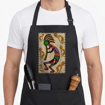 Tablier de cuisine noir avec illustration colorée d'un Kokopelli amérindien entouré de motifs géométriques et de petites figures dansantes.