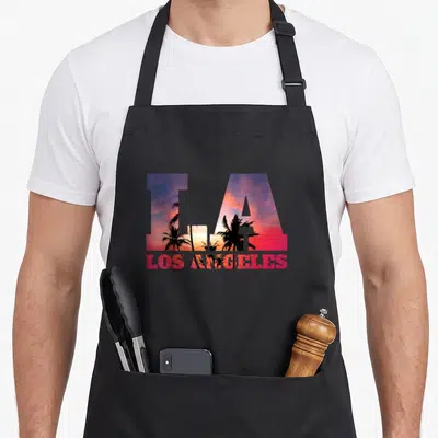 Tablier de cuisine noir avec les lettres LA et LOS ANGELES sur fond de coucher de soleil et palmiers.
