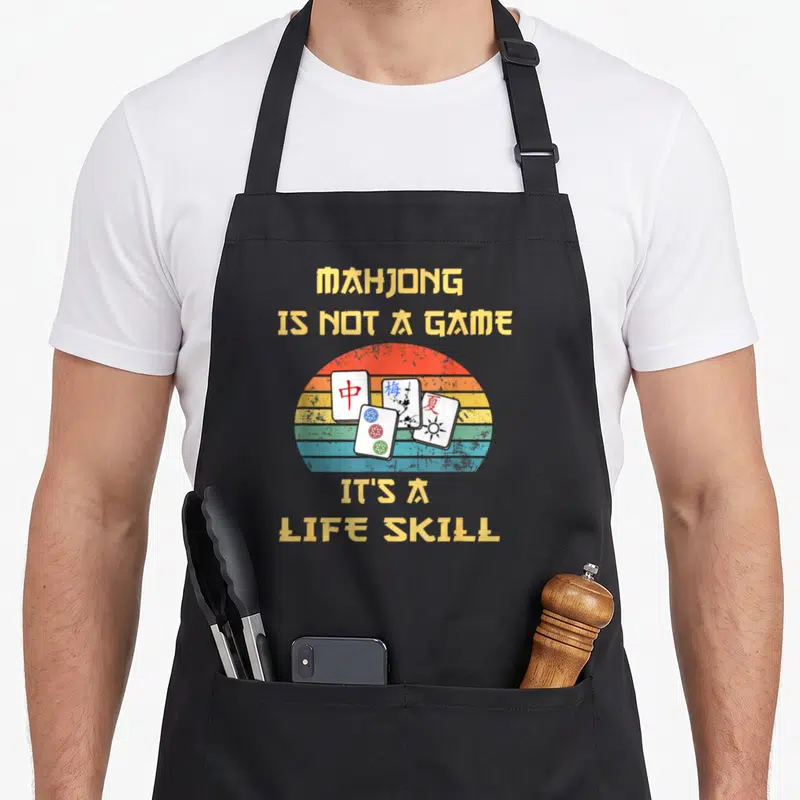 Tablier de cuisine noir avec illustration de tuiles de Mahjong et texte jaune sur fond arc-en-ciel délavé.
