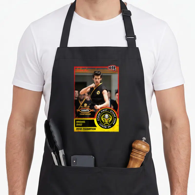 Tablier de cuisine noir avec une illustration de Miguel Diaz en tenue de karaté et le logo Cobra Kai.