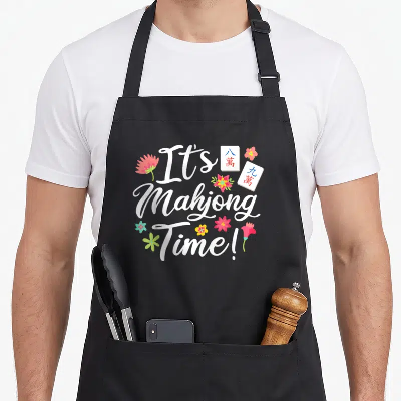 Tablier de cuisine noir avec le texte It's Mahjong Time entouré de tuiles et de fleurs colorées.