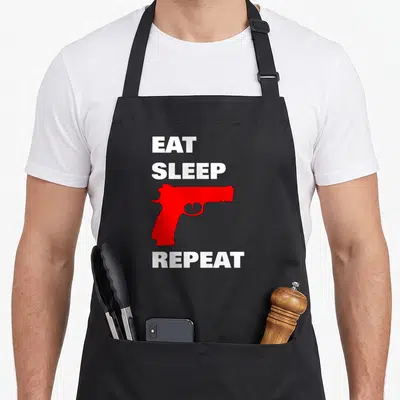 Tablier de cuisine noir avec un pistolet rouge et le texte blanc Eat Sleep Shoot Repeat Freedom.