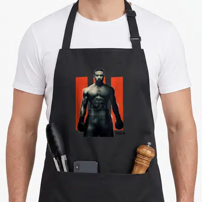 Tablier de cuisine noir avec portrait d'Adonis Creed posé devant des barres rouges verticales.