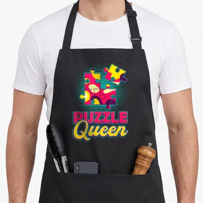 Tablier de cuisine noir avec des pièces de puzzle colorées et le texte Puzzle Queen Puzzling Puzzle imprimé au centre.