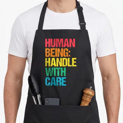 Tablier de cuisine noir avec inscription multicolore Human Being Handle with Care en typographie épaisse et texturée.