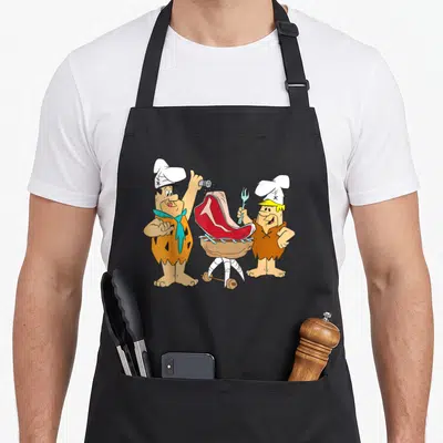 Tablier de cuisine noir avec Fred et Barney en chefs préparant un steak sur un gril en pierre