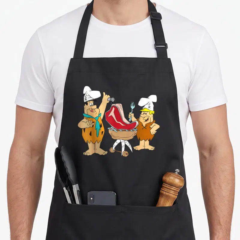 Tablier de cuisine noir avec Fred et Barney en chefs préparant un steak sur un gril en pierre