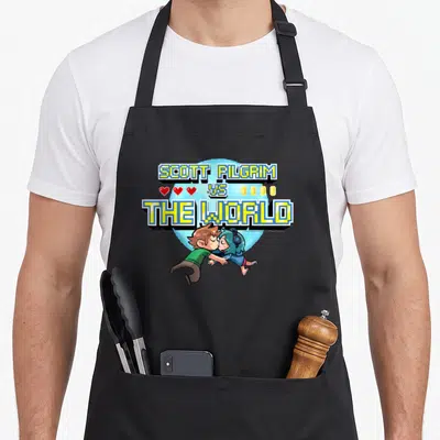 Tablier de cuisine noir avec logo Scott Pilgrim Vs The World et personnages en pixel art colorés sur fond bleu.