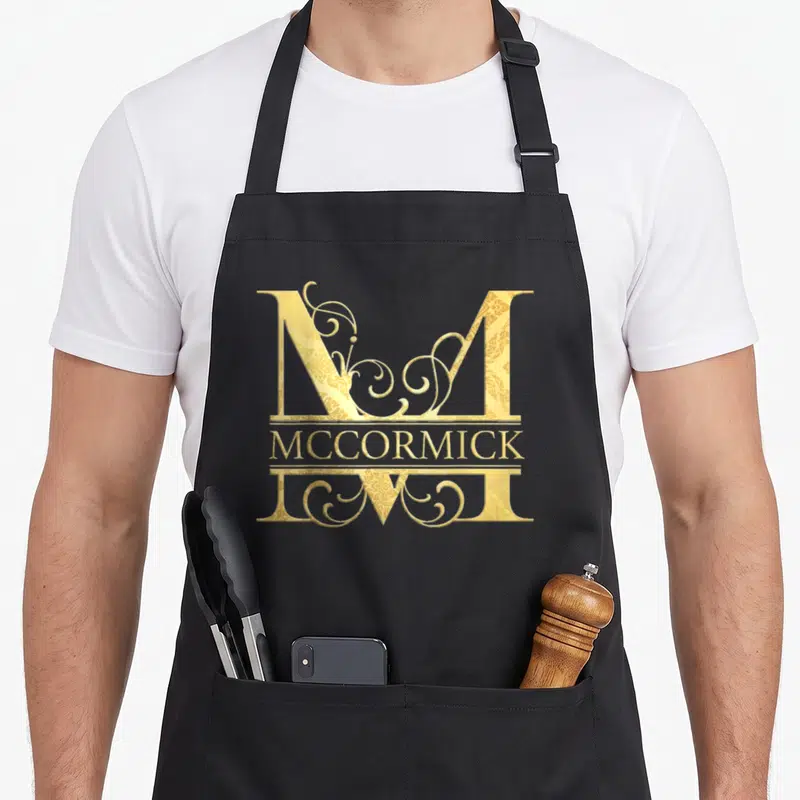 Tablier de cuisine noir avec un grand M doré orné de volutes et le nom McCormick imprimé au centre.