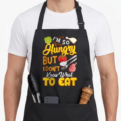 Tablier de cuisine noir avec inscription J'ai tellement faim mais je ne sais pas quoi manger entourée d'aliments et d'ustensiles.