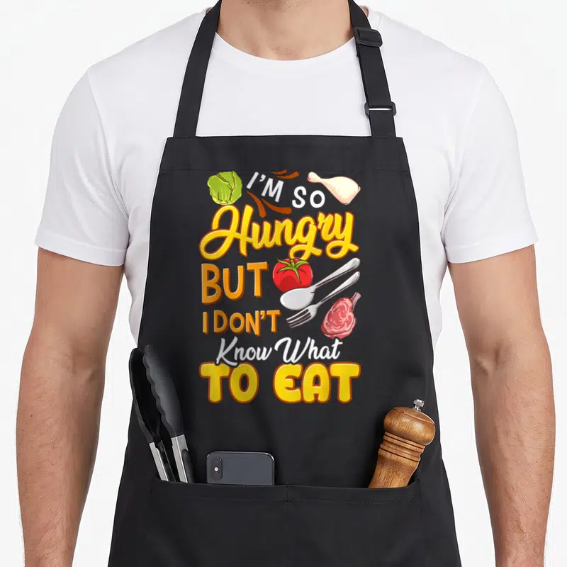 Tablier de cuisine noir avec inscription J'ai tellement faim mais je ne sais pas quoi manger entourée d'aliments et d'ustensiles.