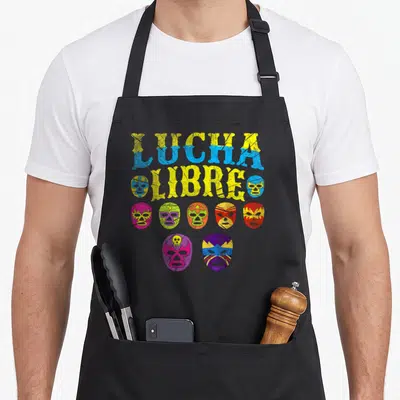 Tablier de cuisine noir avec sept masques de luchador multicolores et l'inscription LUCHA LIBRE en lettres jaunes et bleues texturées.
