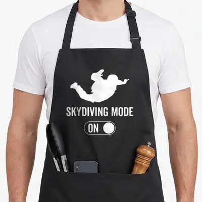 Tablier de cuisine noir avec silhouette blanche d'un parachutiste et texte Skydiving Mode On sur fond uni.