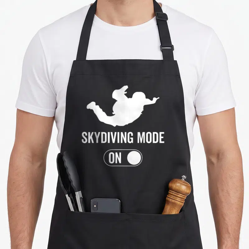 Tablier de cuisine noir avec silhouette blanche d'un parachutiste et texte Skydiving Mode On sur fond uni.