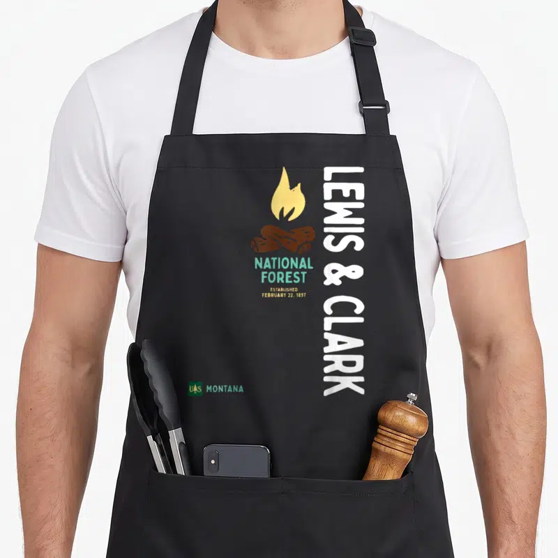 Tablier de cuisine noir avec logo Lewis & Clark National Forest Montana et illustration de feu de camp.