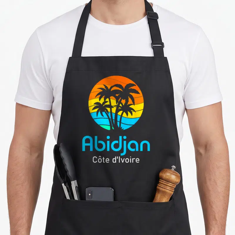 Tablier de cuisine noir avec logo circulaire coloré représentant des palmiers et le texte Abidjan Côte d'Ivoire en bas.