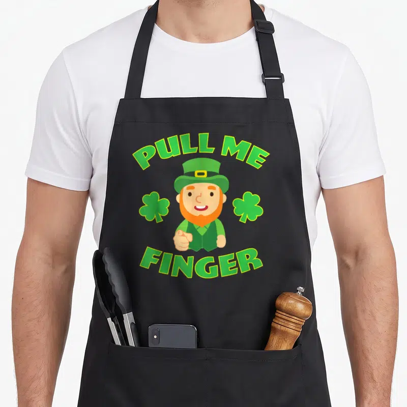 Tablier de cuisine noir avec lutin roux pointant et texte Pull Me Finger encadré de trèfles verts.