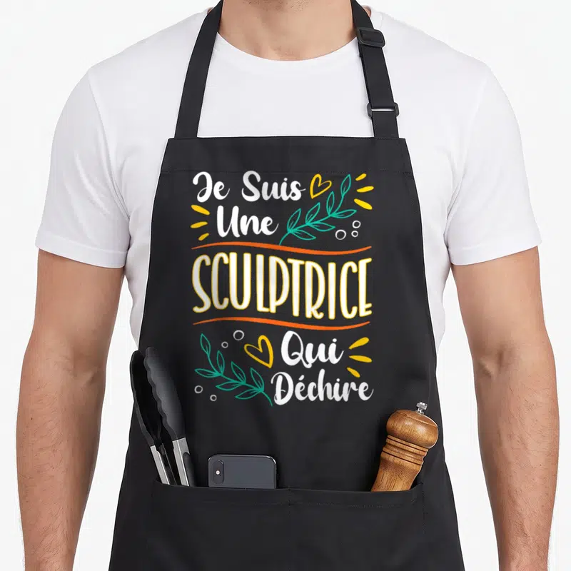 Tablier de cuisine noir avec l'inscription Je Suis Une SCULPTRICE Qui Déchire en blanc et jaune, orné de feuilles vertes et de cœurs oranges.