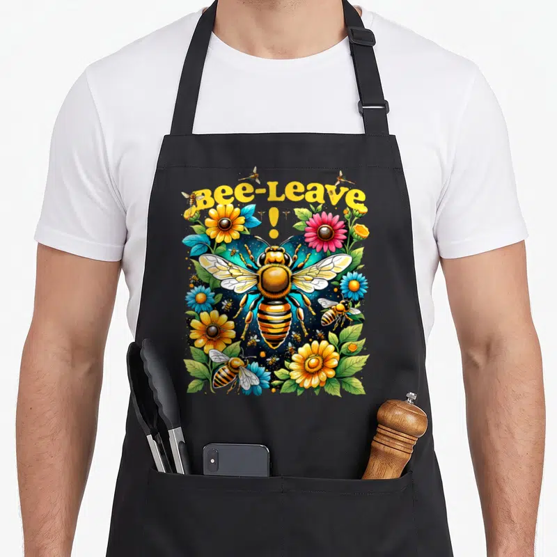 Tablier de cuisine noir avec une grande abeille, des fleurs jaunes et roses, et le texte Bee-Leave. en jaune.