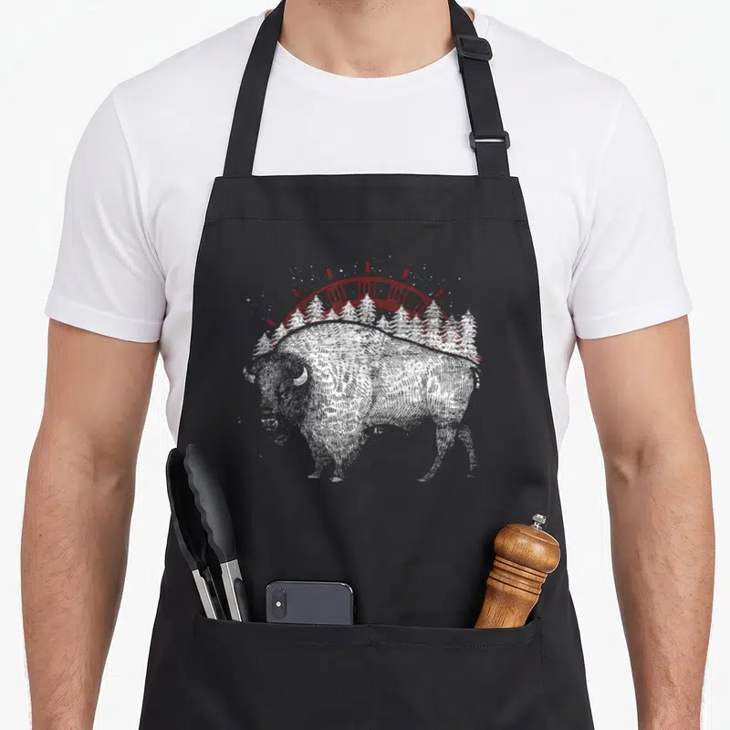 Tablier de cuisine noir avec illustration d'un buffle américain blanc et d'un arc rouge stylisé au-dessus de sapins.