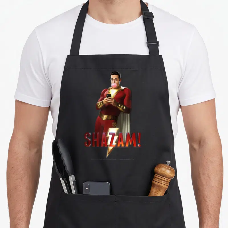 Tablier de cuisine noir avec Shazam. en costume rouge mâchant du bubble gum rose et tenant un téléphone portable.