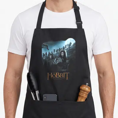 Tablier de cuisine noir avec une aventure de personnages fantastiques et le titre The Hobbit en lettres dorées sur fond bleu nuit.