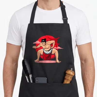 Tablier de cuisine noir avec illustration colorée d'un lutteur de sumo en position de combat devant un soleil rouge et un portail japonais.