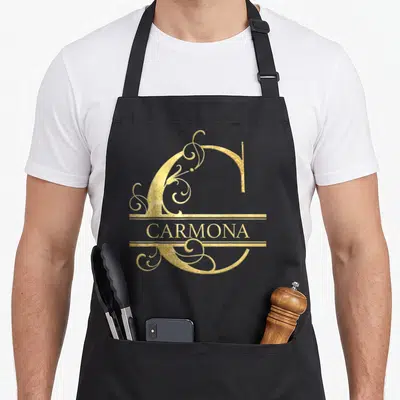 Tablier de cuisine noir avec un grand C doré orné d'arabesques et le nom Carmona en lettres capitales.