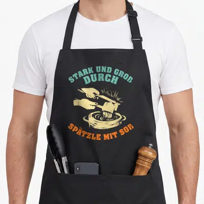 Tablier de cuisine noir avec une illustration de mains préparant du spätzle et du texte en allemand.