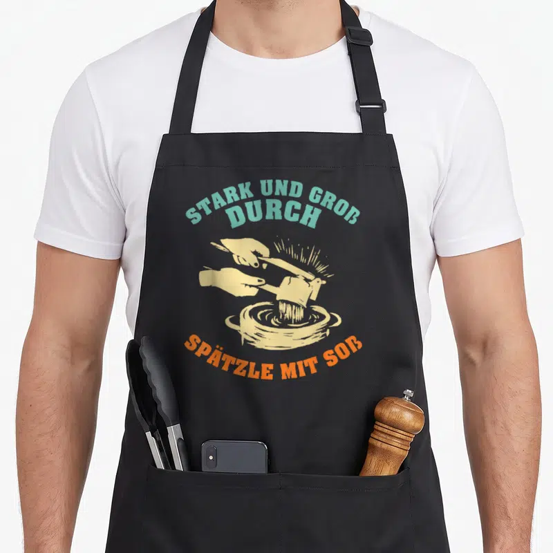 Tablier de cuisine noir avec une illustration de mains préparant du spätzle et du texte en allemand.