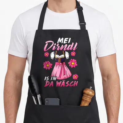 Tablier de cuisine noir avec illustration de dirndl rose et texte Mei Dirndl is in da Wäsch entouré de fleurs.