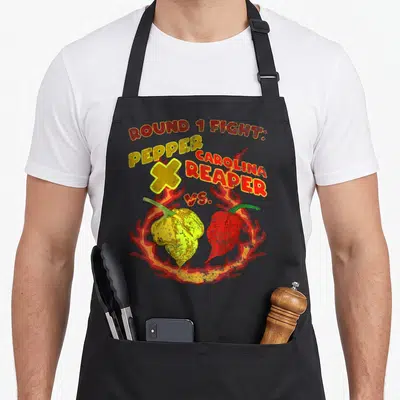 Tablier de cuisine noir avec illustration de duel entre Pepper X et Carolina Reaper entourés de flammes.