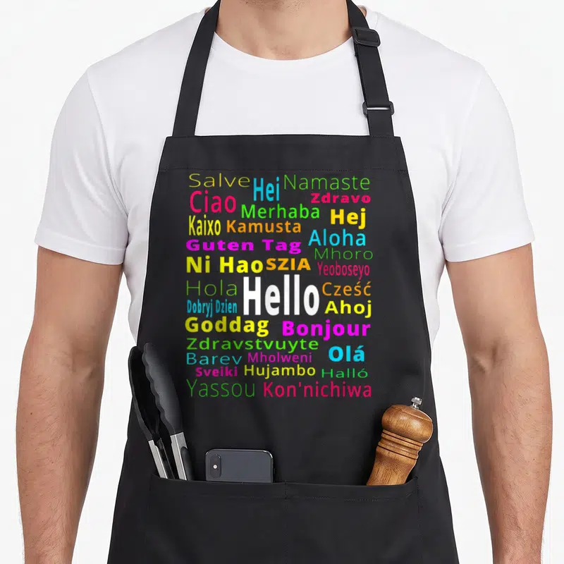 Tablier de cuisine noir avec Hello, Bonjour et d'autres salutations multicolores disposées en nuage typographique.