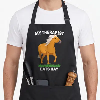 Tablier de cuisine noir avec illustration d'un cheval Haflinger orange et texte blanc My Therapist Eats Hay sur fond gris uni.