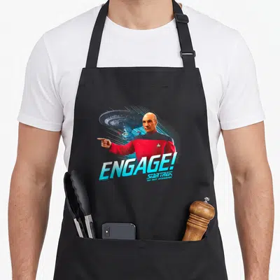 Tablier de cuisine noir avec le capitaine Picard pointant et le texte ENGAGE en bleu sous la navette spatiale.