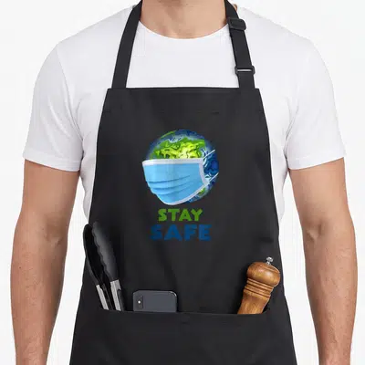 Tablier de cuisine noir avec globe terrestre portant un masque chirurgical bleu et le texte stay safe imprimé en vert et bleu.