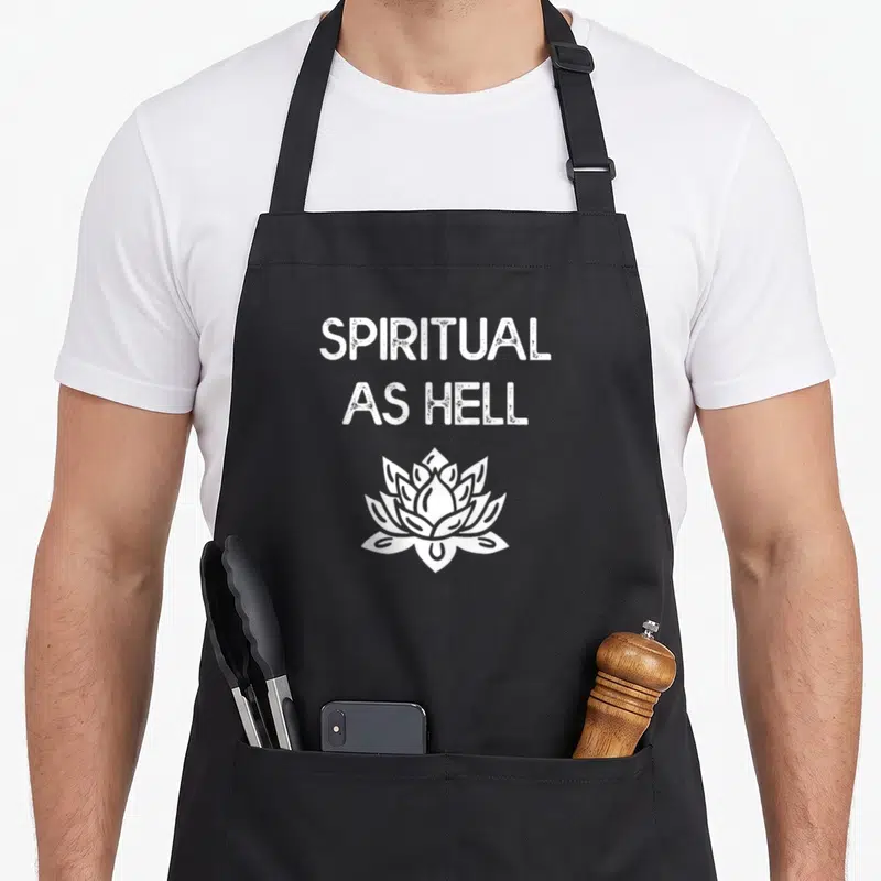 Tablier de cuisine noir avec l'inscription Spiritual As Hell et une fleur de lotus blanche stylisée.