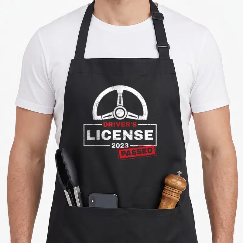Tablier de cuisine noir avec volant blanc et inscription Driver's « License » 2023 Passed en rouge et blanc.