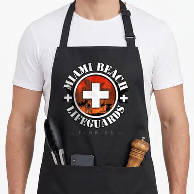 Tablier de cuisine noir avec logo circulaire Miami Beach Lifeguards Florida et croix blanche sur fond orange.