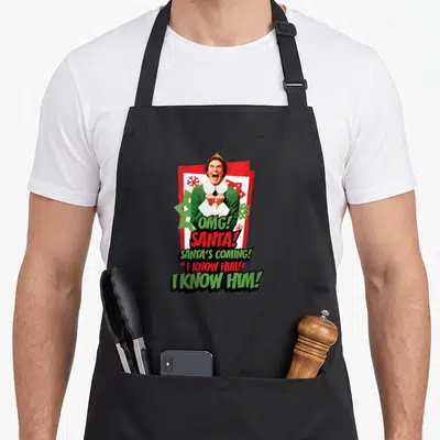 Tablier de cuisine noir avec un elfe criant et le texte « OMG! SANTA! I KNOW HIM! » sur fond de cartes rouges.