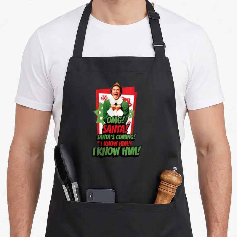 Tablier de cuisine noir avec un elfe criant et le texte « OMG! SANTA! I KNOW HIM! » sur fond de cartes rouges.