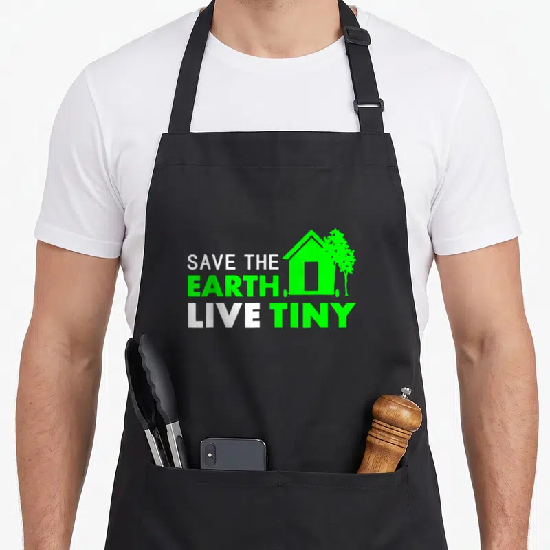 Tablier de cuisine noir avec le texte Save the Earth Live Tiny House et une petite maison verte stylisée à côté d'un arbre.
